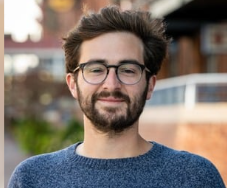 A young man with glasses and beard, wearing a blue sweater, and smiling at the camera. 