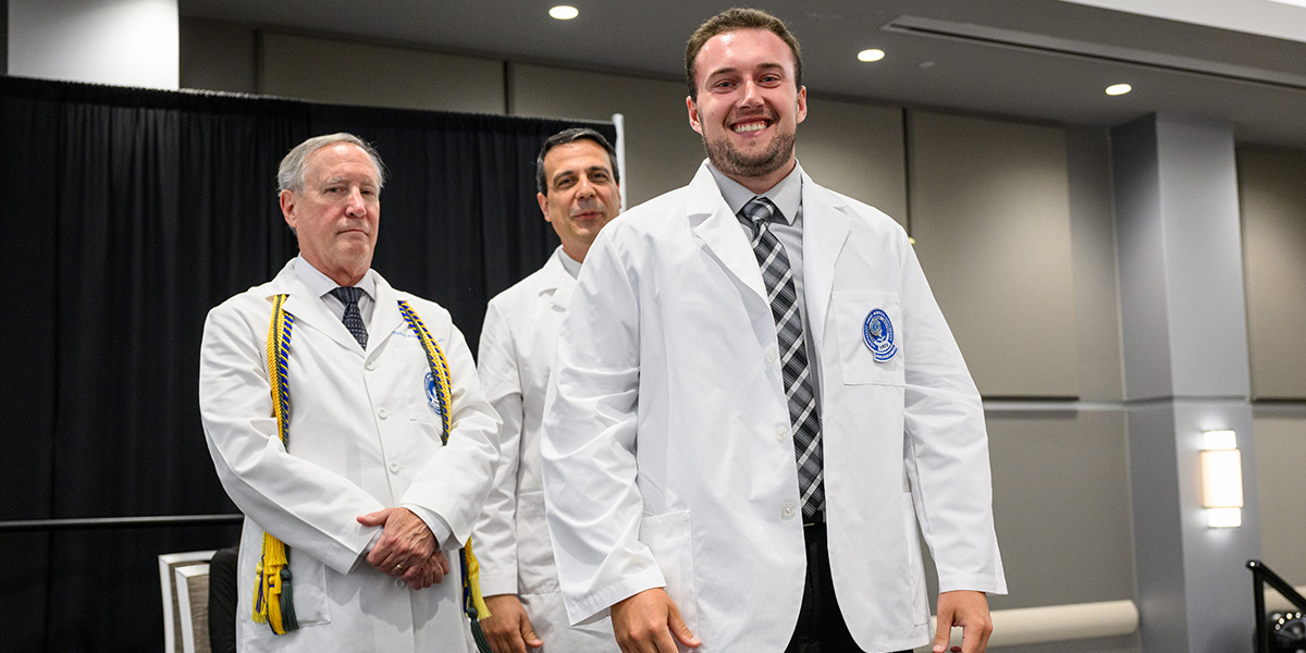 Young man shows off his new white coat while two faculty members look on