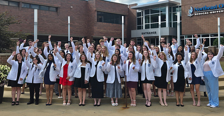 Large group of pharmacy students in white coats pump their fists in celebration