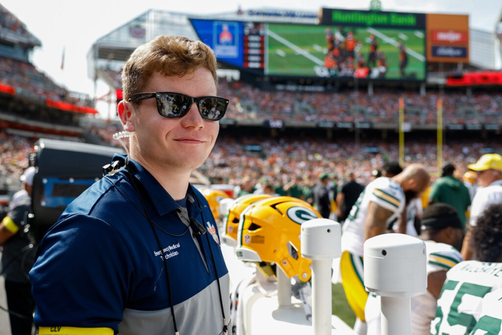 Man wearing sunglasses, jacket, and bookbag smiling while standing at the Browns stadium