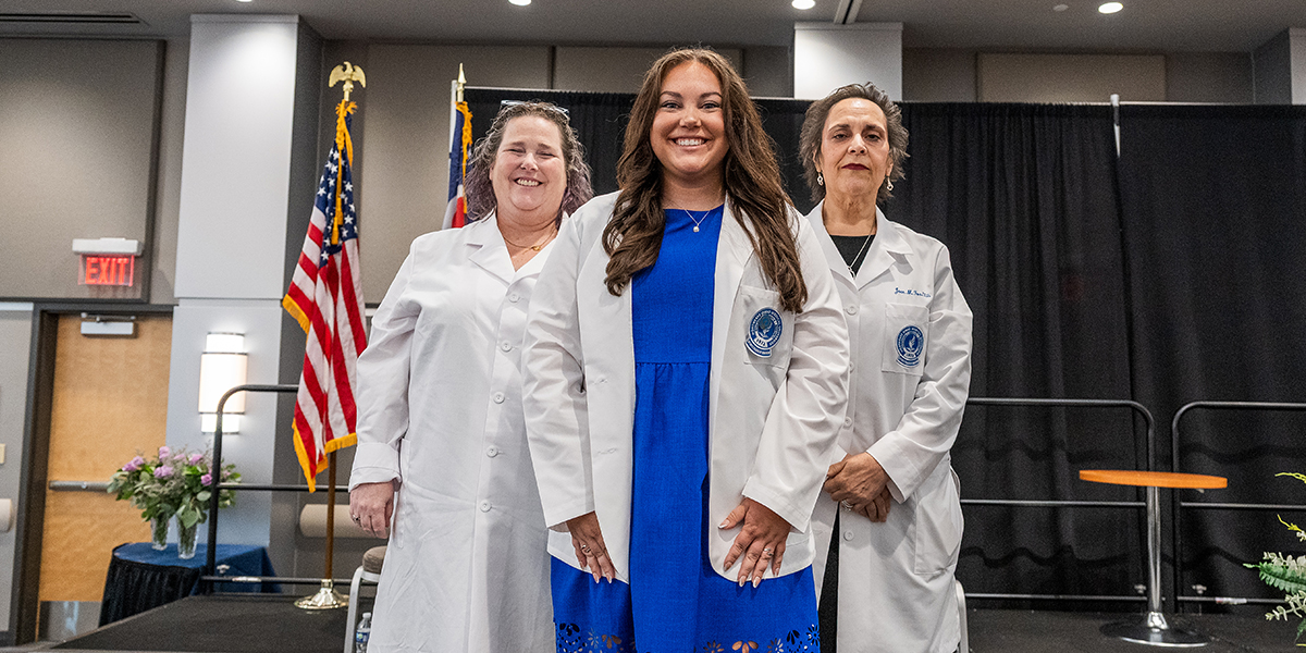 Young woman in blue dress stands on stage with her new white coat between two faculty members