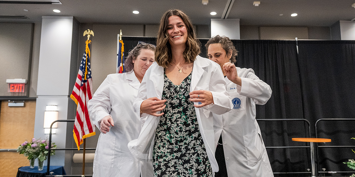 Young woman in a patterned dress receives her white coat on stage from two faculty members