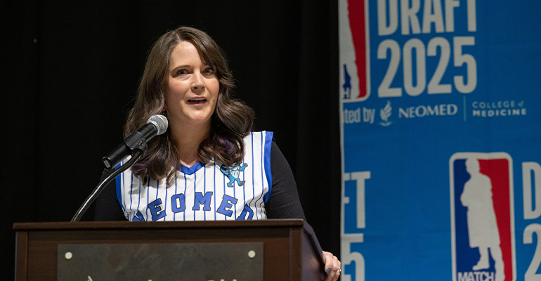 Woman wearing basketball jersey speaks at a podium standing in front of a "Draft 2025" sign