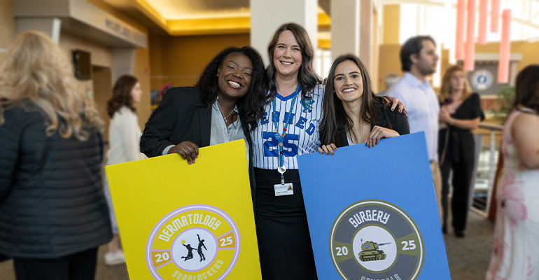A faculty member stands with two graduating medical students who are holding signs related to Match Day.