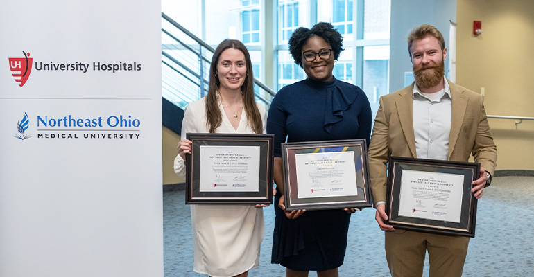 Three PhD students stand side-by-side holding framed certificares next to a UH-NEOMED pop-up banner