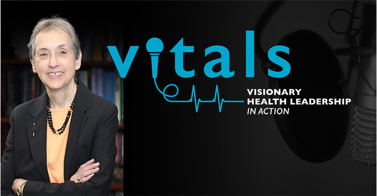 Nina Schor stands with arms folded in front of black background including VITALS logo