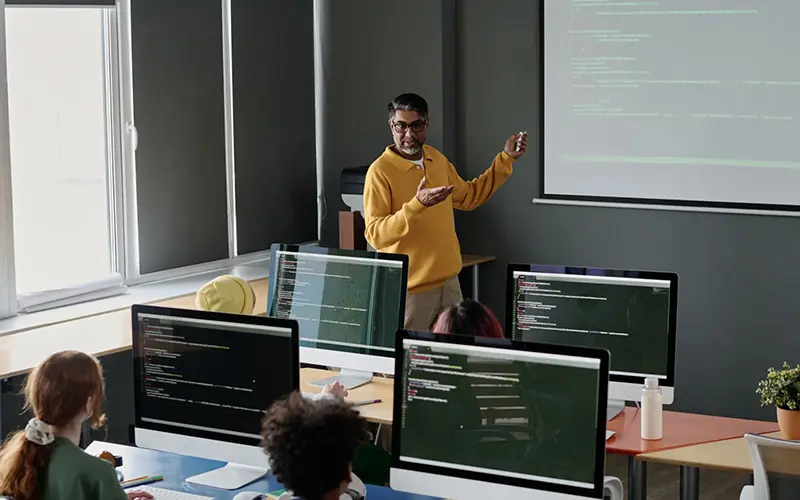 A teacher in a yellow sweater gestures toward a projection screen displaying code while students at desktop computers follow along in a data science and analytics classroom.