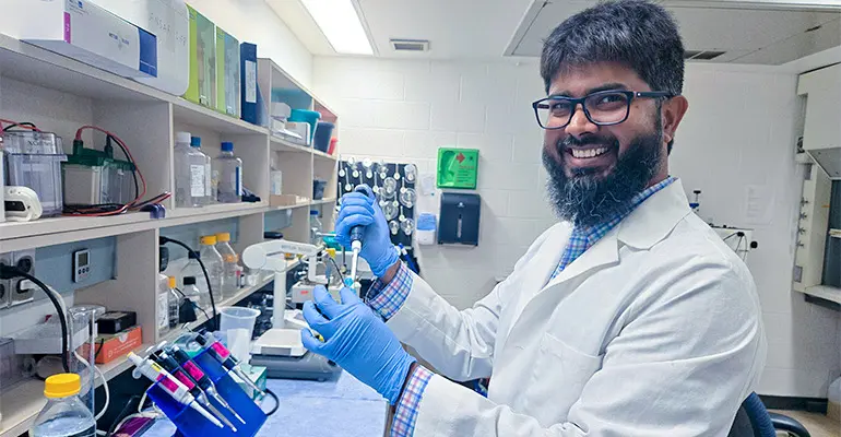 A researcher wearing a lab coat and gloves smiles while using a pipette at a laboratory bench filled with scientific equipment and supplies.