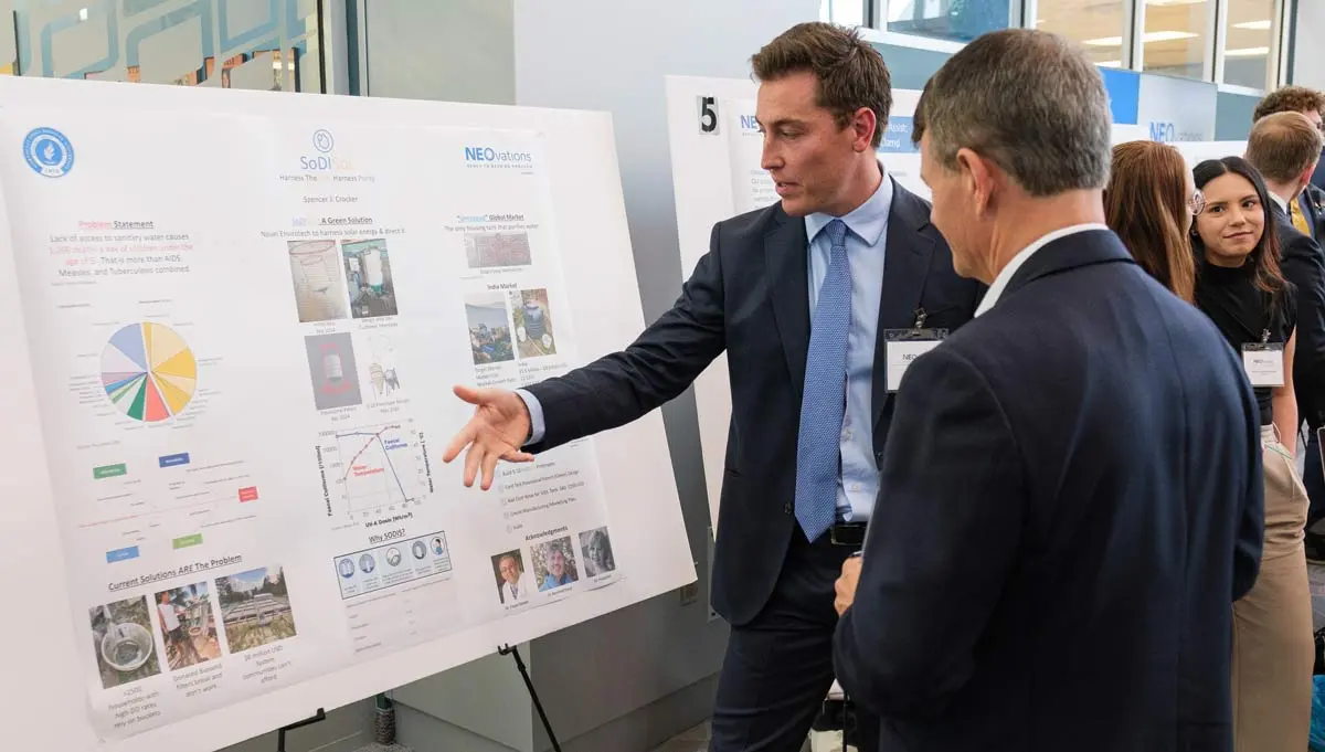 A medical student in a coat and tie explains his medical innovation to another man.