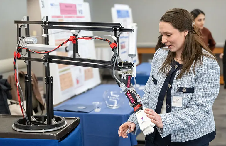 An entrepreneur demonstrates a mechanical arm prototype at NEOMED’s Bench to Bedside innovation showcase, standing beside display boards and equipment on a table.