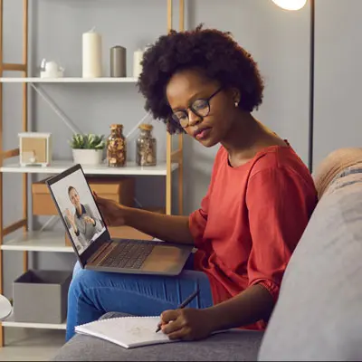 A mental-health professional takes notes while participating in a virtual training session on her laptop from a comfortable home setting.