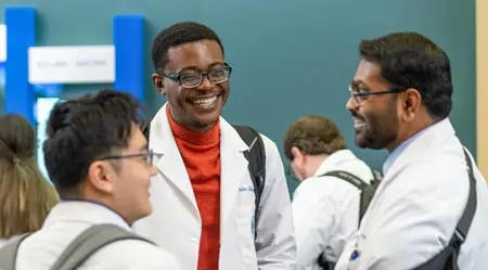Three students in white coats share a laugh during a social event on campus