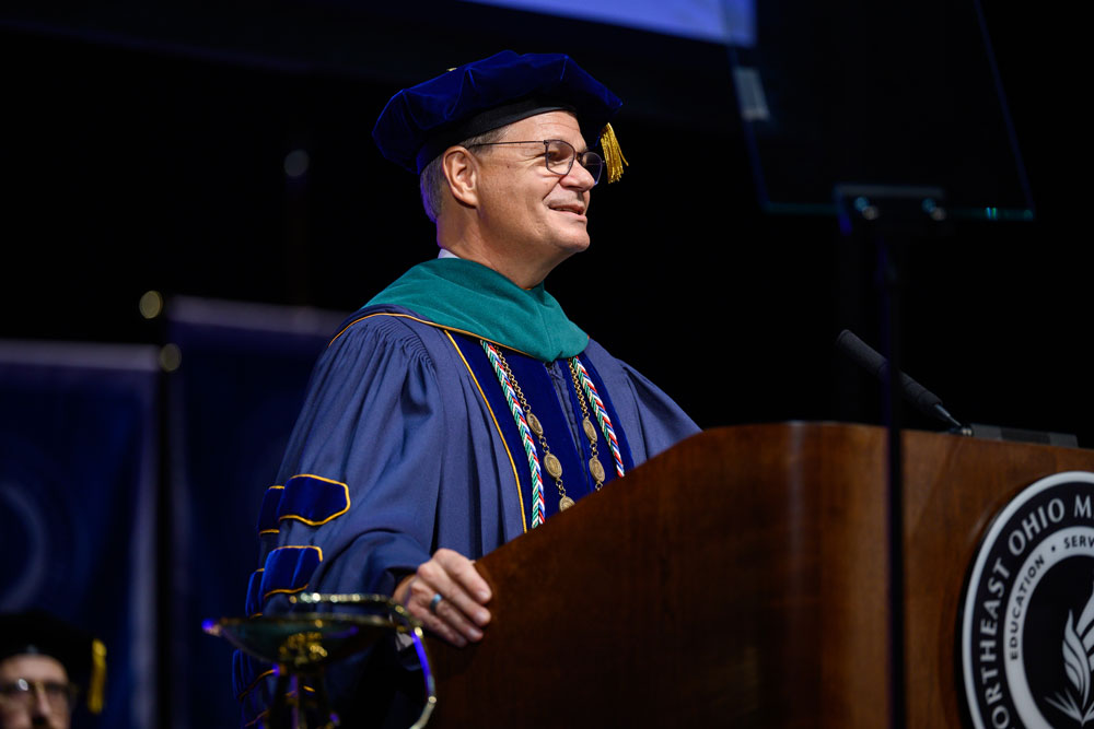 University President Dr. John Langell in academic regalia speaks at a Northeast Ohio Medical University podium while faculty members sit on stage behind him.