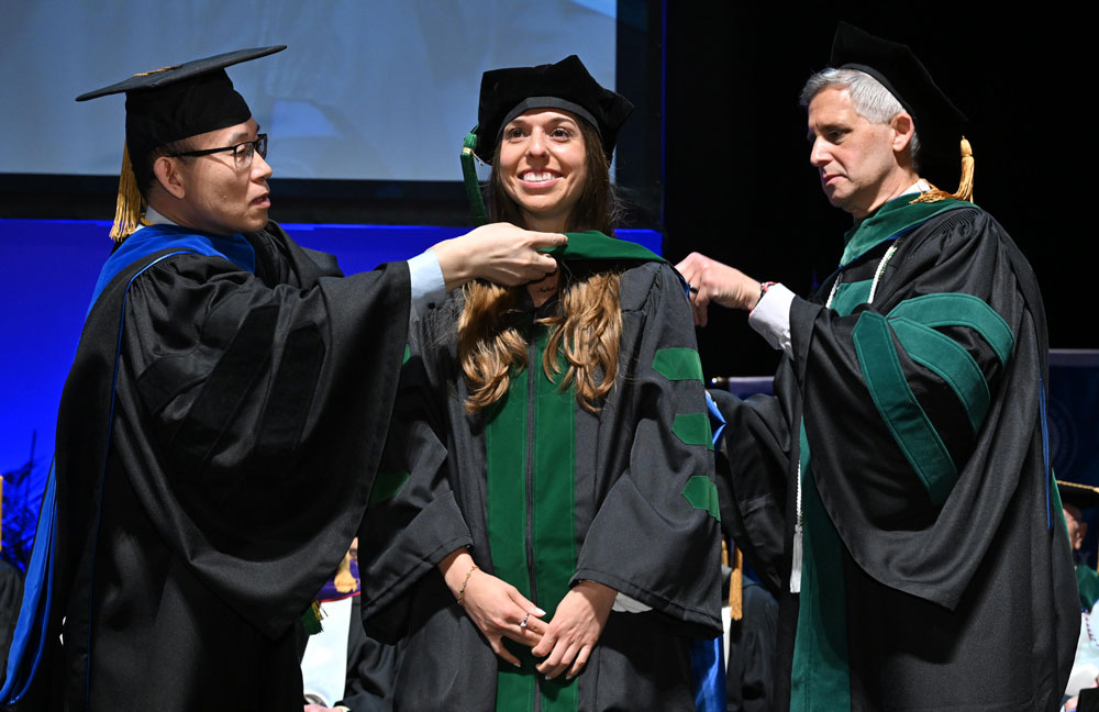 Two faculty members place a doctoral hood on a graduate during a commencement ceremony.
