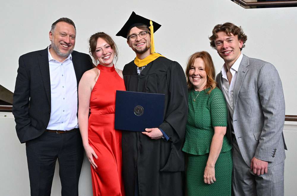A graduate in cap and gown holds a diploma cover while posing with four guests after commencement.