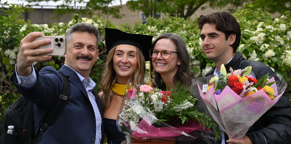 A graduate in cap and gown poses outdoors with three guests while holding flowers after commencement.