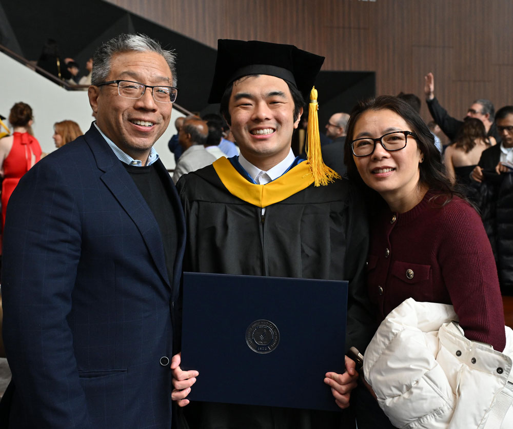 A graduate in cap and gown smiles while holding a diploma cover and posing with two guests at a commencement reception.