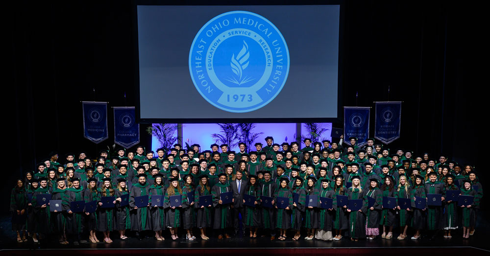 Graduates in academic regalia pose together on stage beneath the Northeast Ohio Medical University seal during commencement.