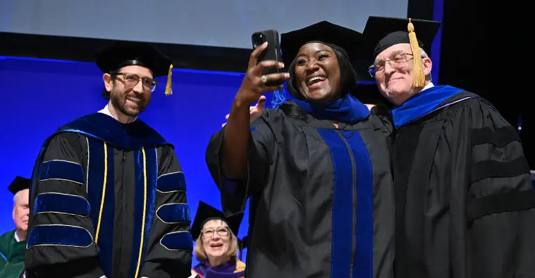 A graduate in academic regalia smiles while taking a selfie with two faculty members on stage during commencement.