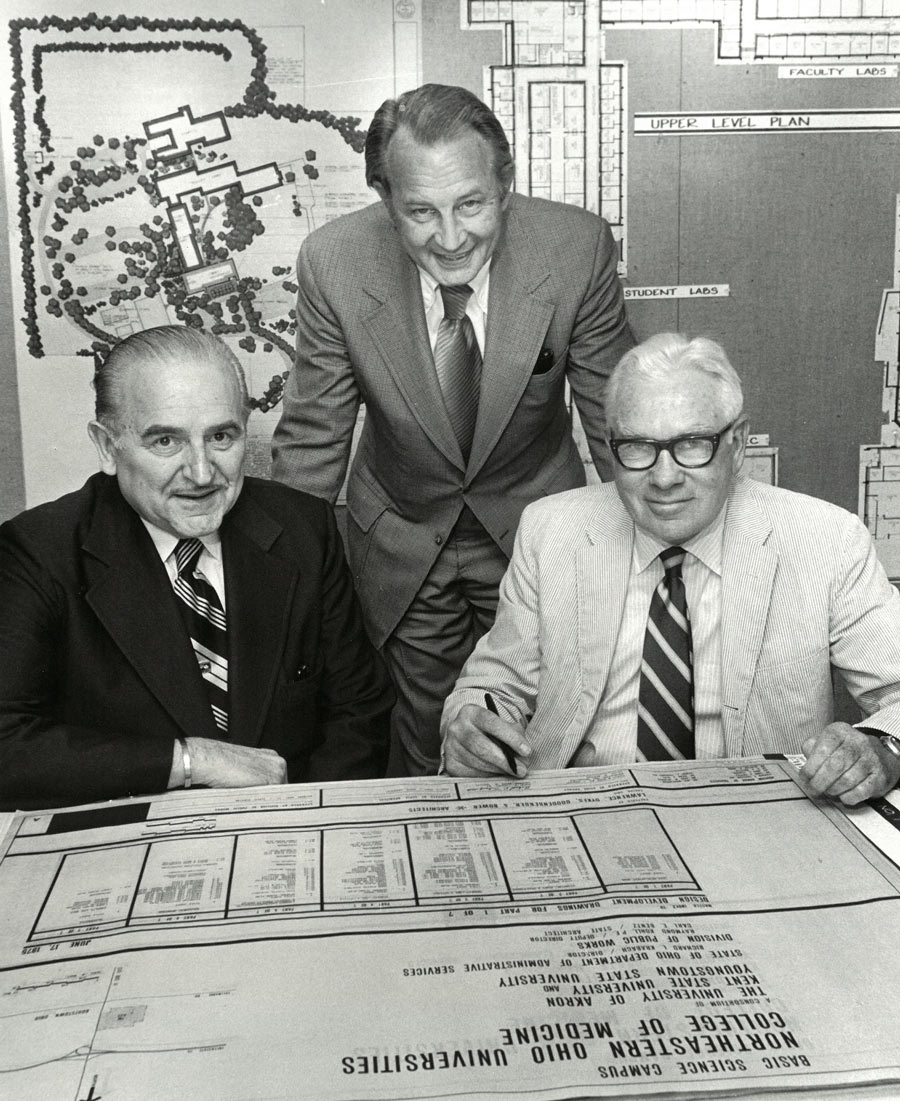 Three men gather around architectural plans, with campus and building diagrams displayed behind them.