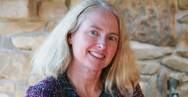 A woman with light-colored hair smiles while standing in front of a stone wall.