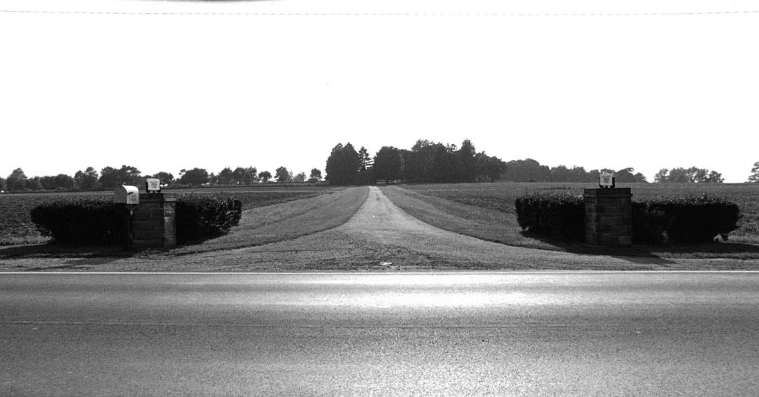 A long driveway extends from a rural road through open fields toward a distant farmhouse surrounded by trees.