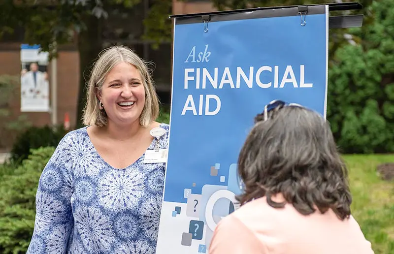 A staff member smiles while speaking with a visitor at an outdoor table beside a sign that reads ‘Ask Financial Aid.’