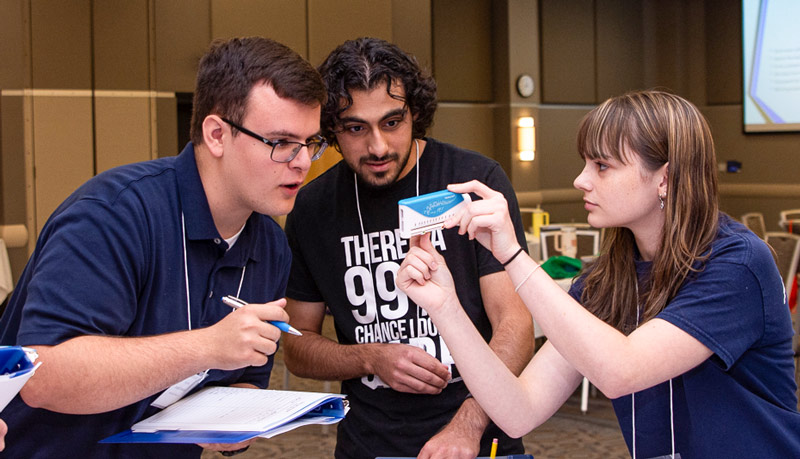Three Health Camp students work together indoors while examining and discussing a small handheld object during an activity.