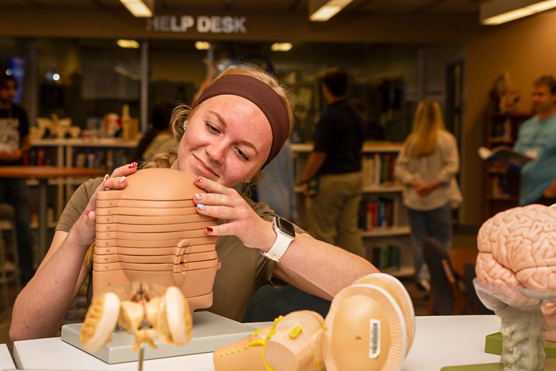 A Health Camp participant adjusts a plastic anatomical model of the human head at a table inside a library or learning space.