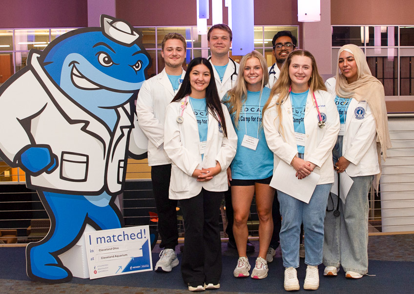 A group of Health Camp students wearing white coats pose indoors beside a large cartoon of NEOMED's mascot, a whale.