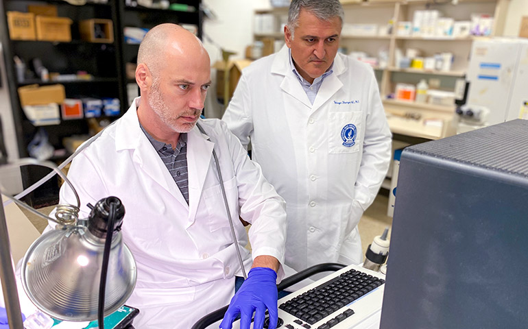 Two researchers in white coats analyze a lab report on a monitor.