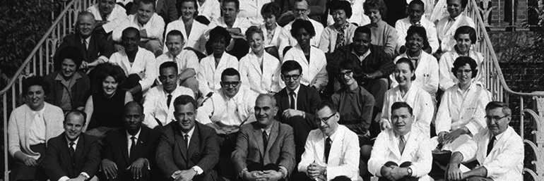 The image is old, in black and white, showing doctors and others sitting on the steps of an ornate building. Most if not all the people are white.