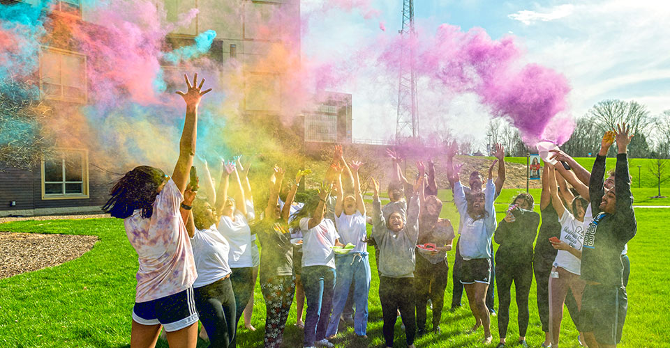 Students throw color dye into the air outside on a sunny day.