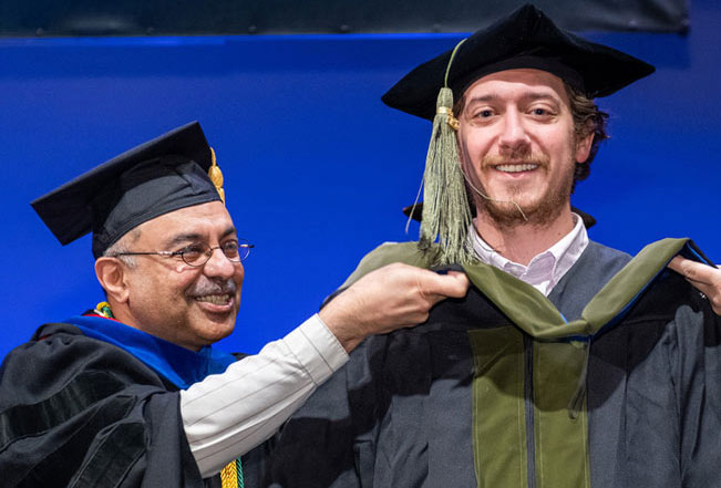 A smiling faculty member places a hood on a student during commencement.