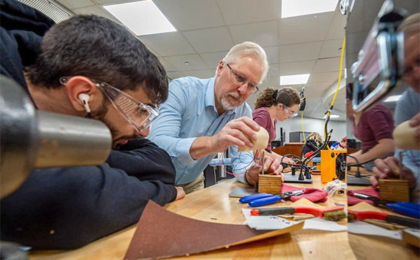 An instructor works closely with students at a lab bench, guiding them as they assemble or test a small prototype using tools and electronic components.