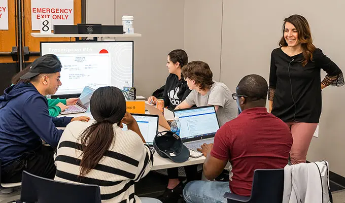 A faculty member talks with students seated around a table as the students work on a problem together.
