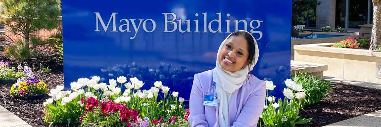A woman sitting outside a sign at the Mayo Clinic, with flowers nearby.