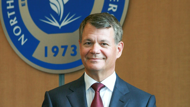 A man in a suit and red tie stands smiling in front of the University seal that includes the year 1973.