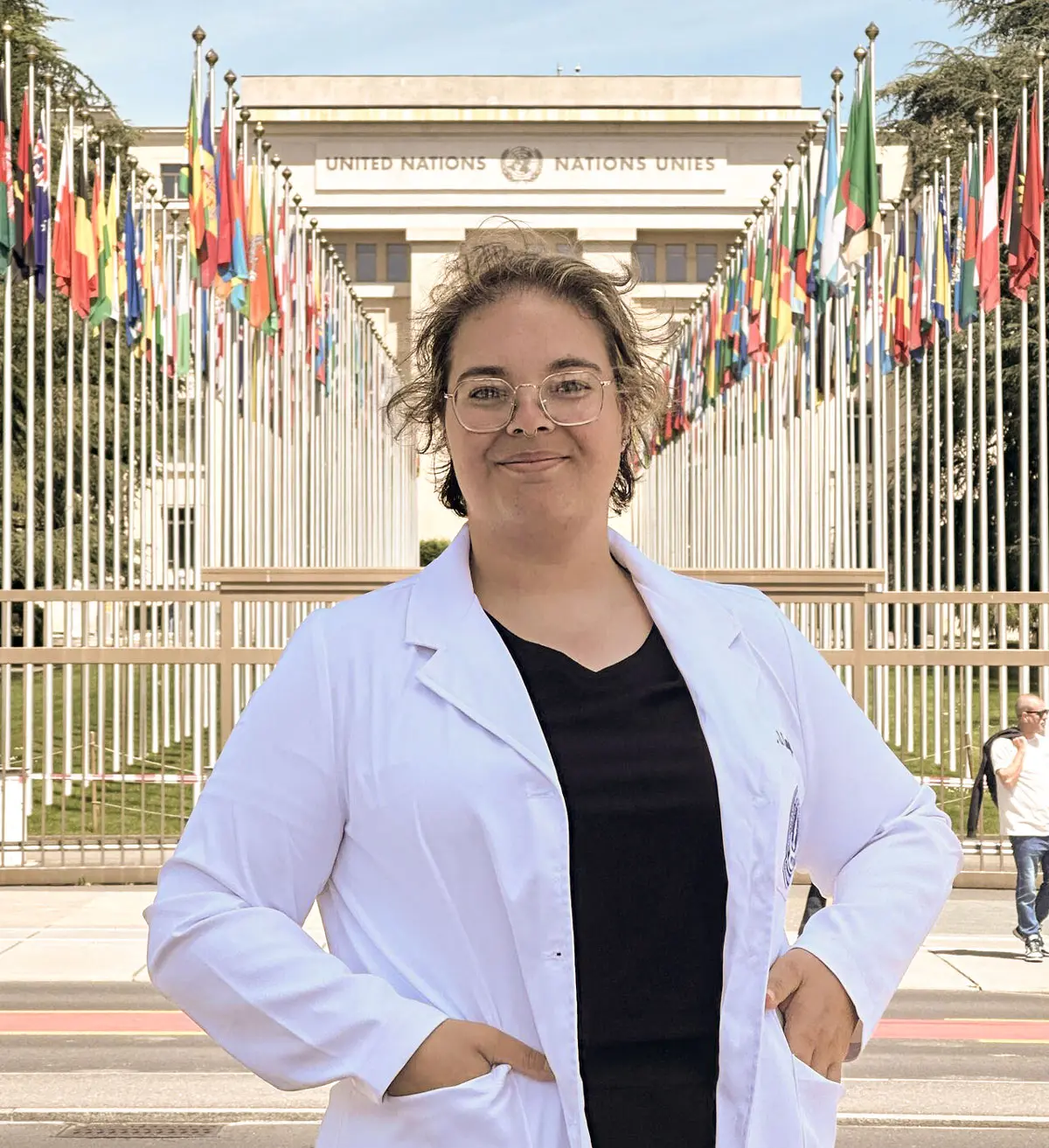 A person wearing a white coat stands confidently in front of the United Nations building, with rows of international flags lining a walkway behind them.