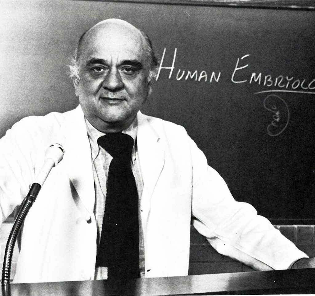 Dr. Liebelt, in a white coat and tie, stands before a chalkboard.