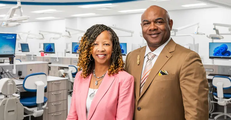 Darrell L. McNair and Ronna J. McNair, in business attire, stand together in NEOMED’s dental simulation lab with workstations and monitors in the background.
