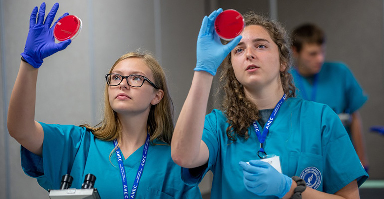 two young females in blue scrubs examine red substance in petri dishes