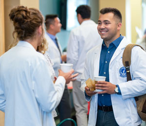 Two medicine students in white coats chat while holding cups of coffee.