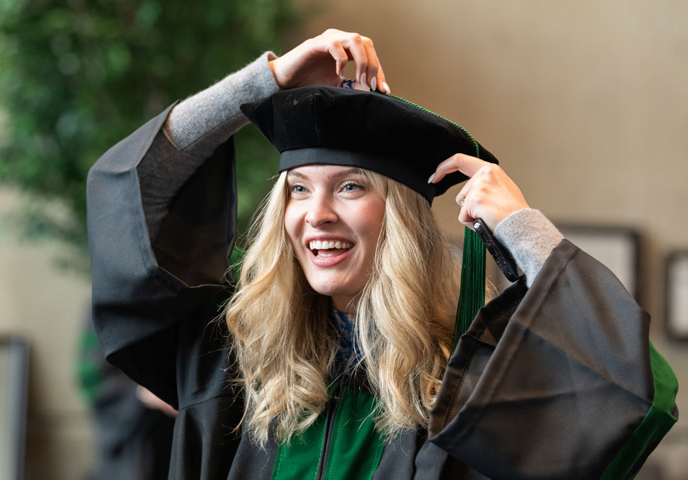A smiling female graduate in doctoral regalia adjusts her tam during commencement preparations, her green-accented robe visible beneath long hair.