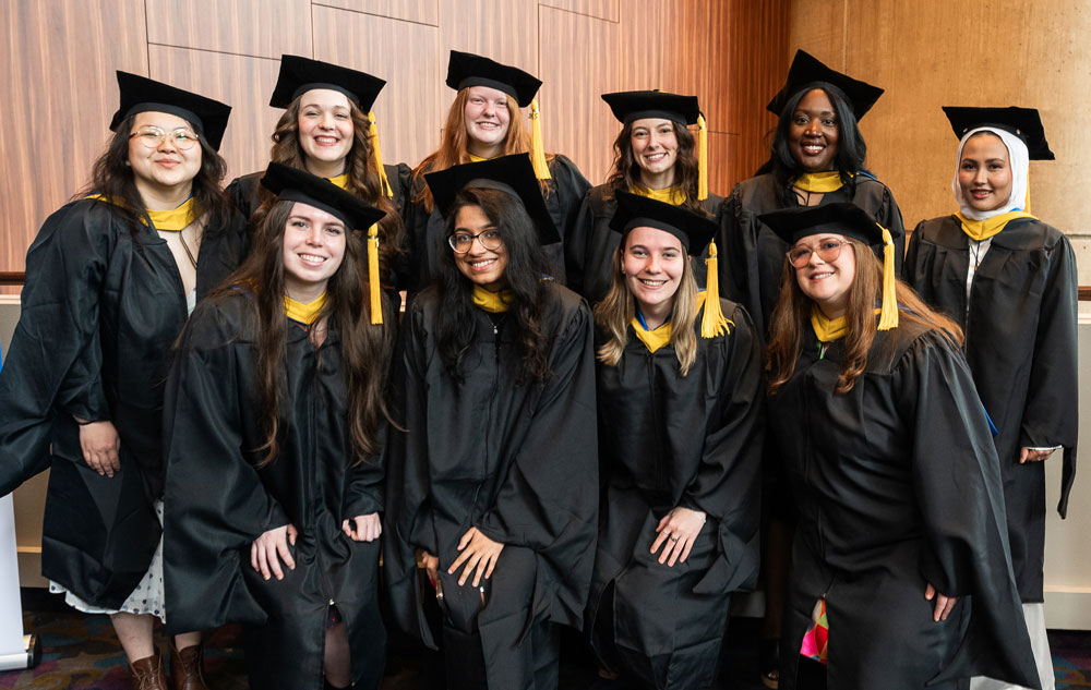 A group of ten female graduates wearing black caps and gowns with yellow stoles pose cheerfully for a photo indoors before their commencement ceremony.