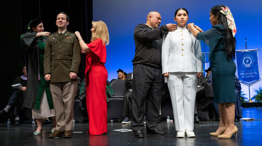 Two uniformed graduates stand at attention while being ceremonially pinned on stage by two women during a commencement recognition for military service.