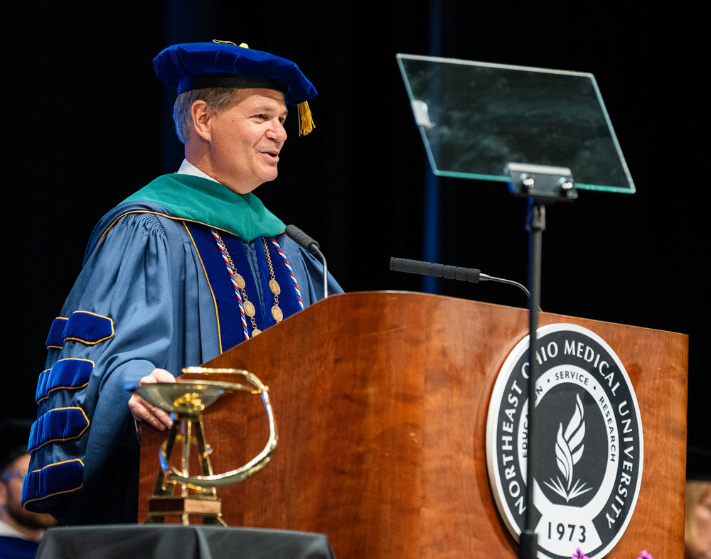 The president of Northeast Ohio Medical University, dressed in blue and green academic regalia, speaks at the podium during the commencement ceremony.