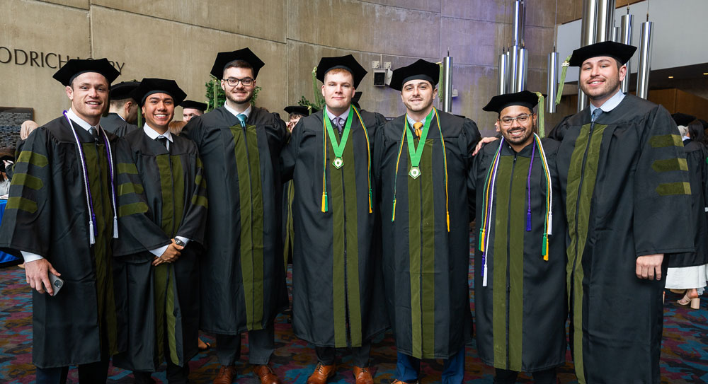 A group of seven male medical school graduates in doctoral regalia, including green-trimmed robes and tams, pose and smile for a photo at a commencement ceremony.