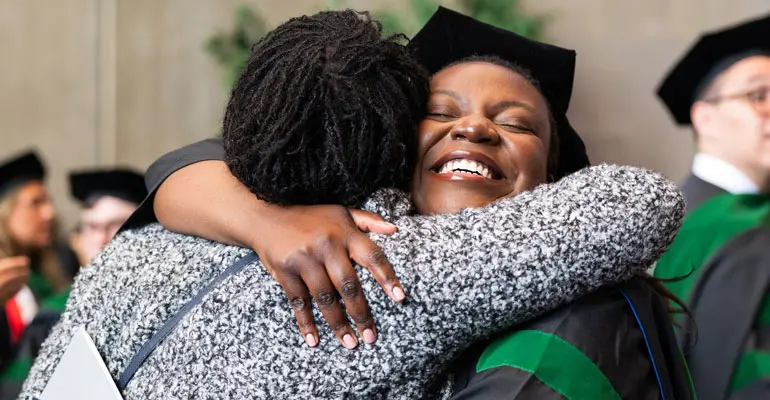 A joyful graduate in doctoral regalia embraces a supporter in a warm hug, her eyes closed and smiling widely during a moment of celebration at commencement.
