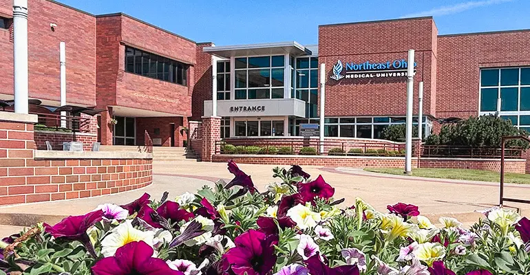 Exterior view of a brick university building with a glass entrance labeled “Entrance,” landscaped flower beds in the foreground, and the Northeast Ohio Medical University sign visible on the facade.
